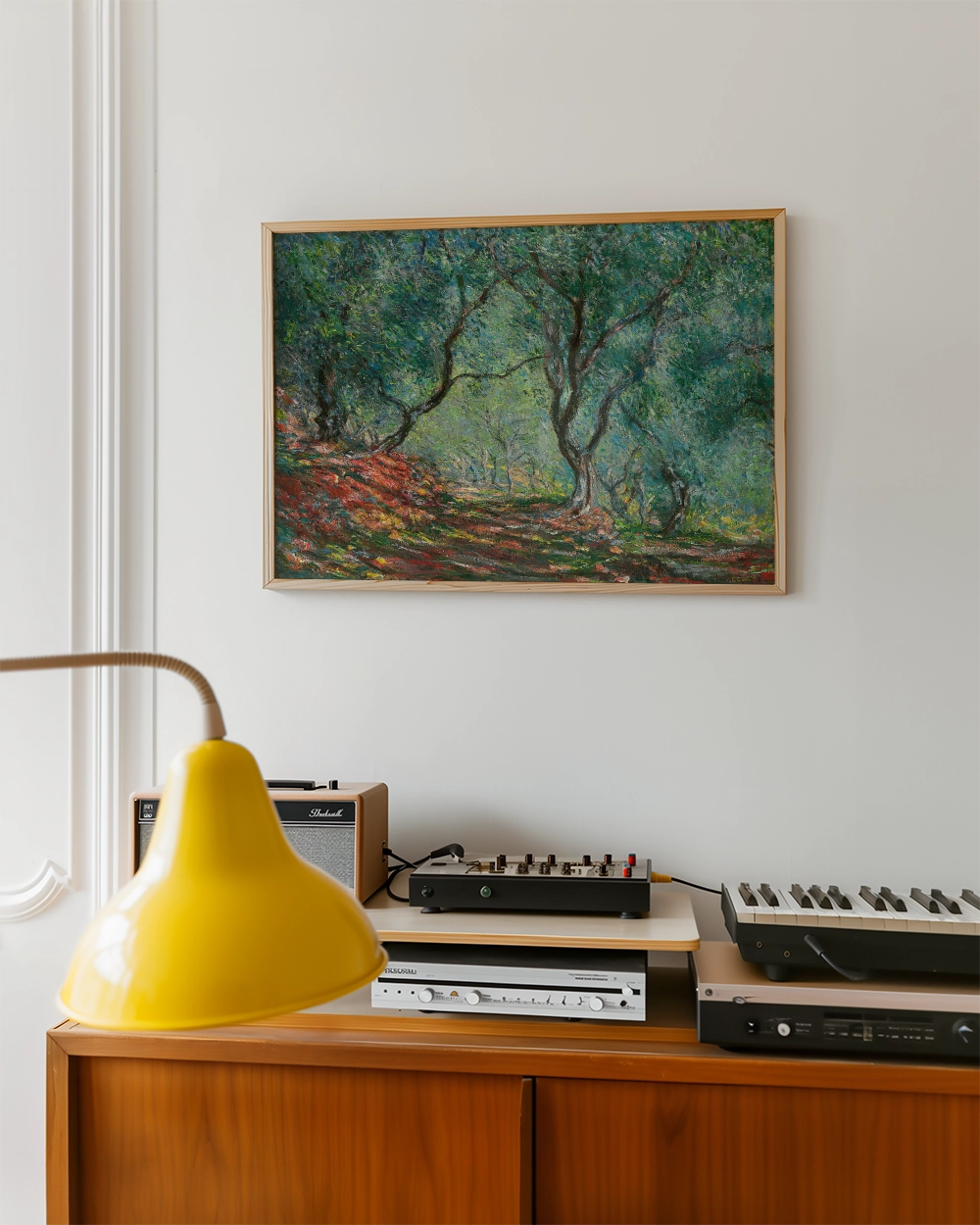Painting of a lush green landscape with trees, framed and displayed on a wall above a wooden console with a yellow lamp and audio equipment.