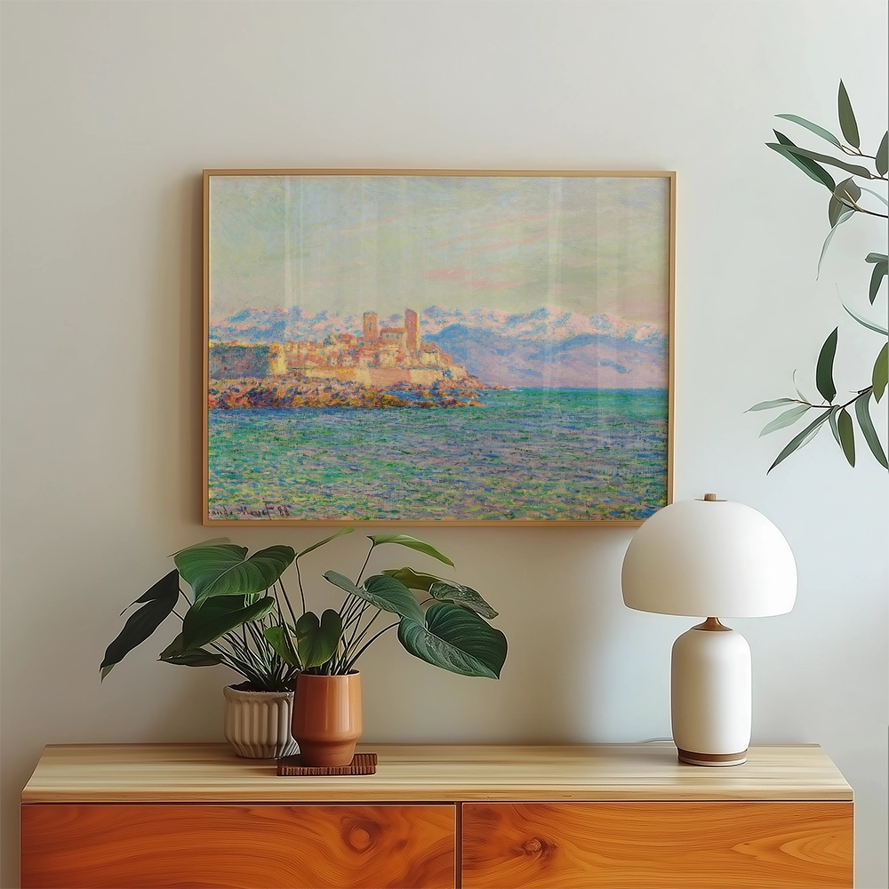 A serene coastal painting depicting a castle on a rocky island surrounded by blue waters and mountains, framed and displayed above a wooden side table with potted plants and a modern lamp.