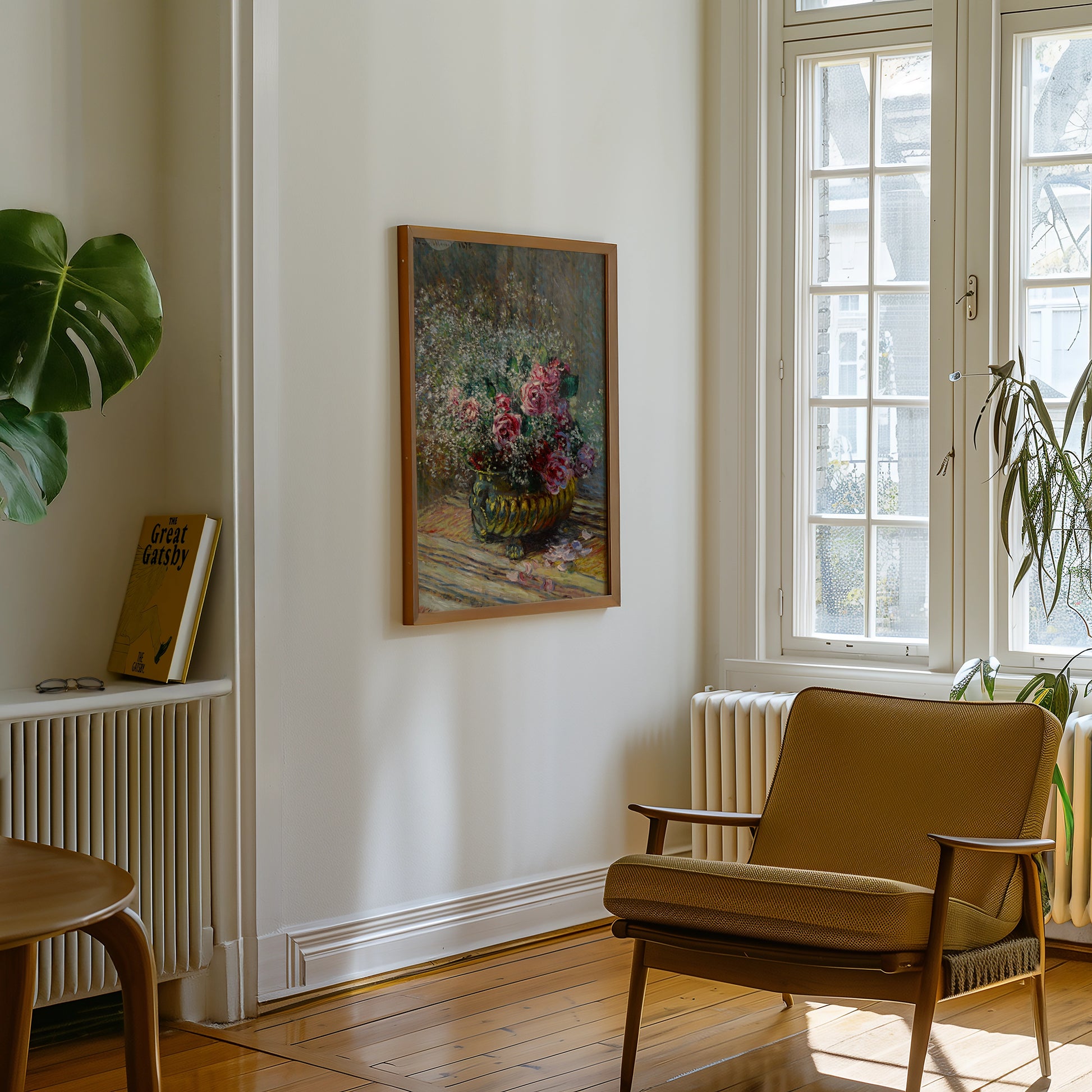 Painting of a flower vase with pink roses hanging on a wall in a bright, modern living room with a brown armchair and a houseplant.