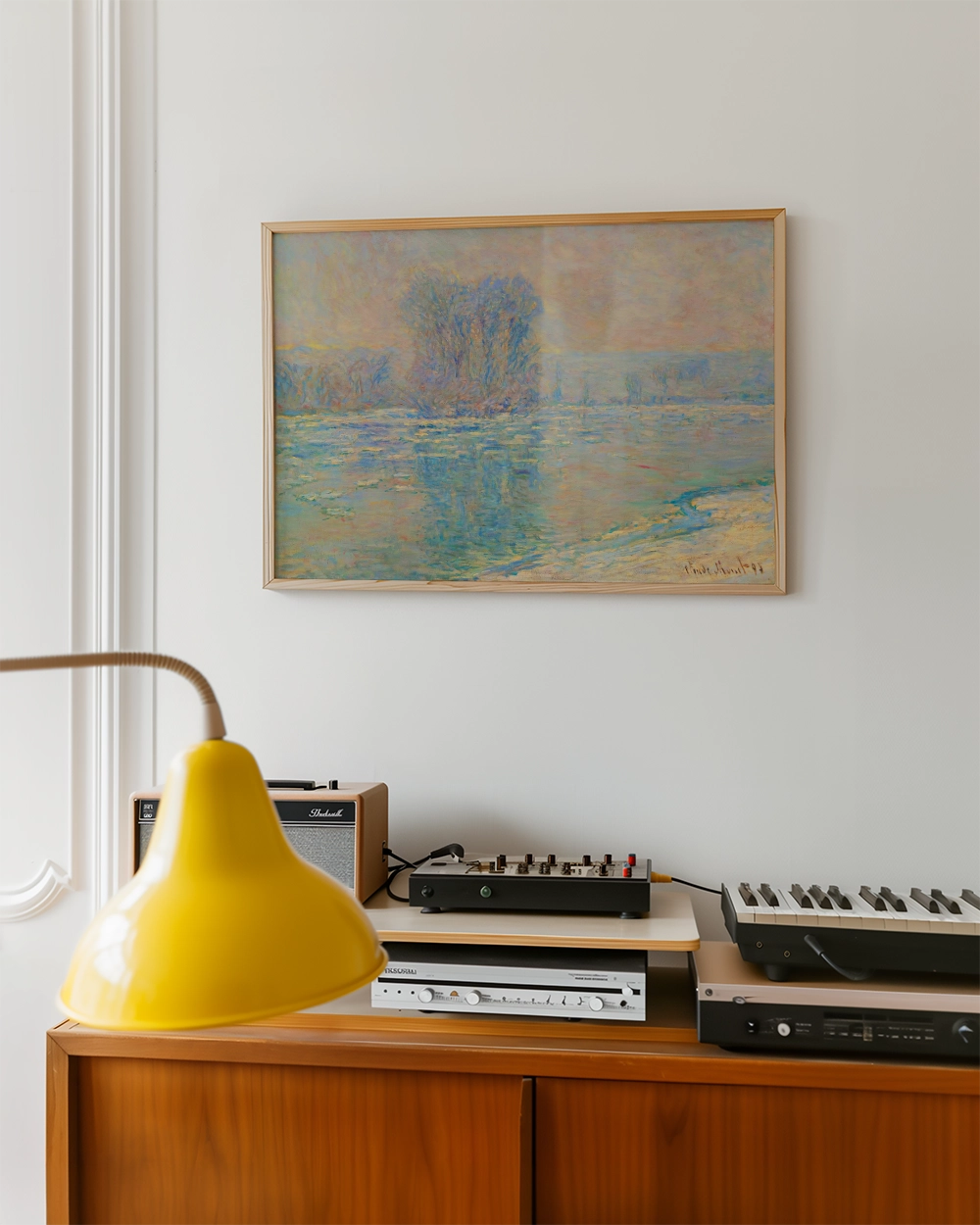 A framed painting of a serene landscape with trees reflected in water, displayed on a white wall above a wooden cabinet featuring a yellow lamp and audio equipment.