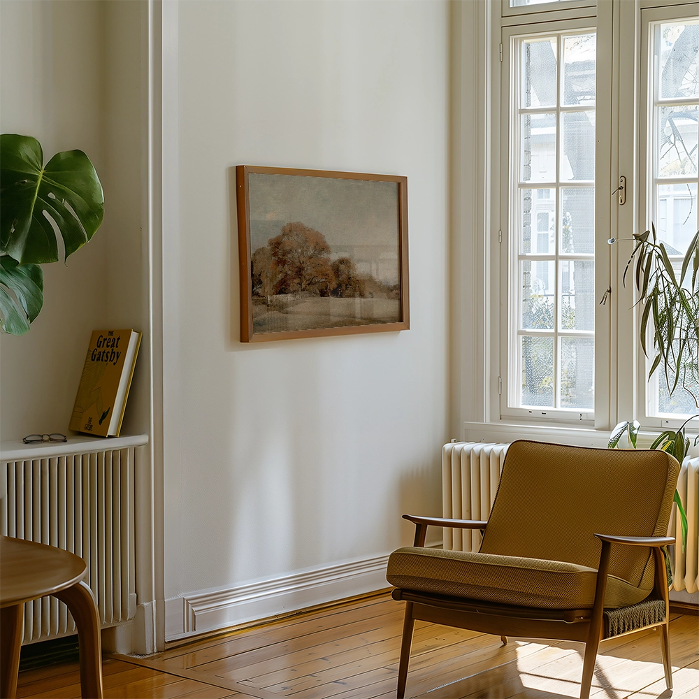 Framed landscape painting hanging on a white wall in a cozy living room setting with a chair and potted plant.