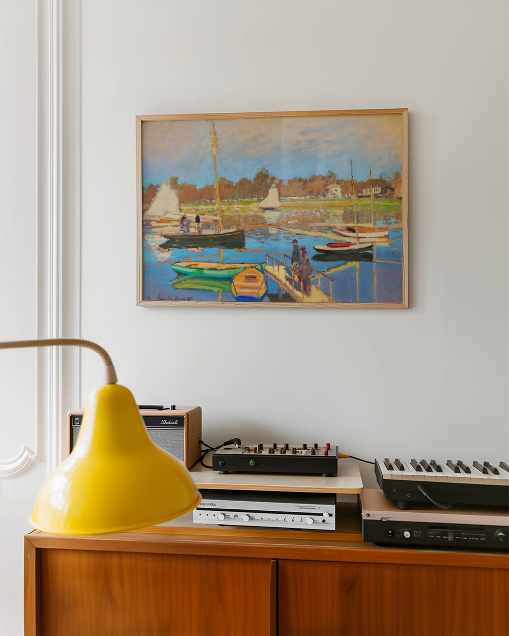 Painting of boats and people by a dock, framed and hung on a wall above a wooden cabinet with a yellow lamp and audio equipment.