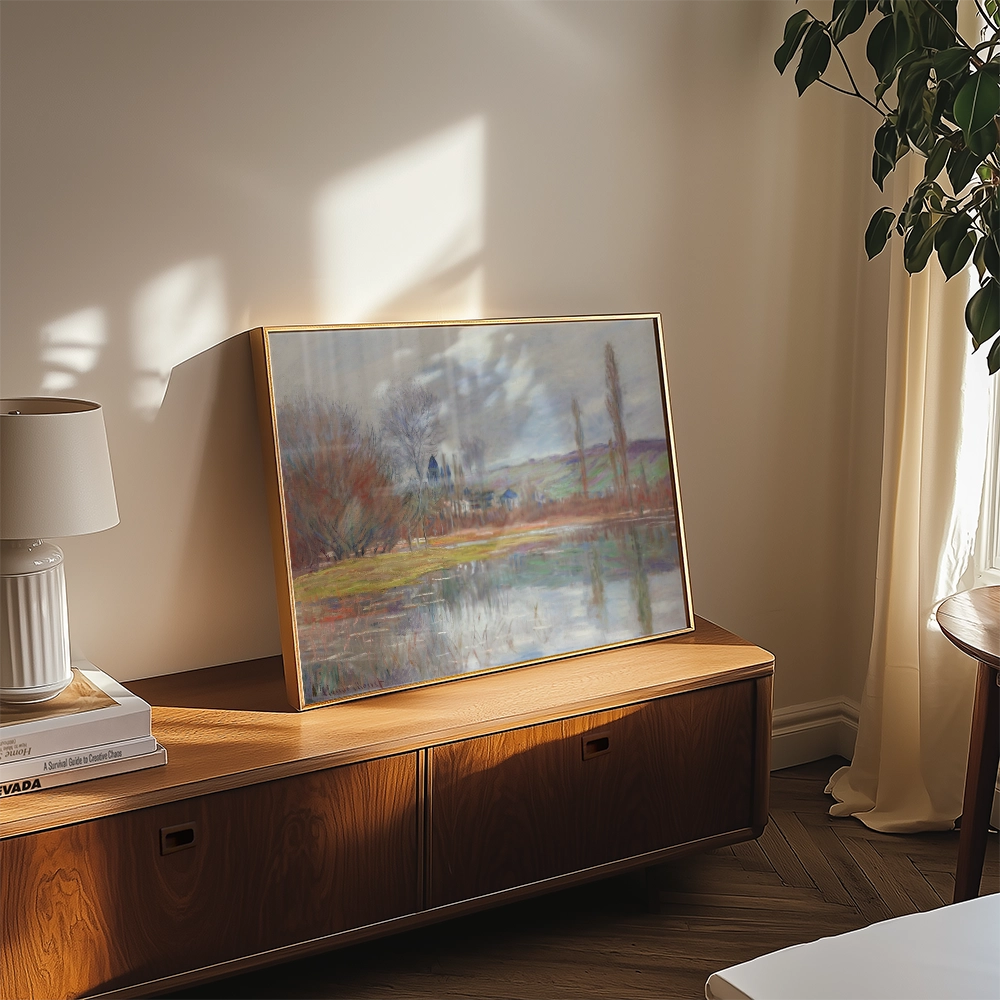 A framed watercolor landscape painting displaying a serene lake and trees, placed on a wooden side table next to a lamp.