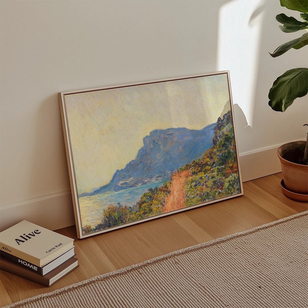 Painting of a scenic landscape with mountains and a path, framed and leaning against a wall, with books and a plant nearby.