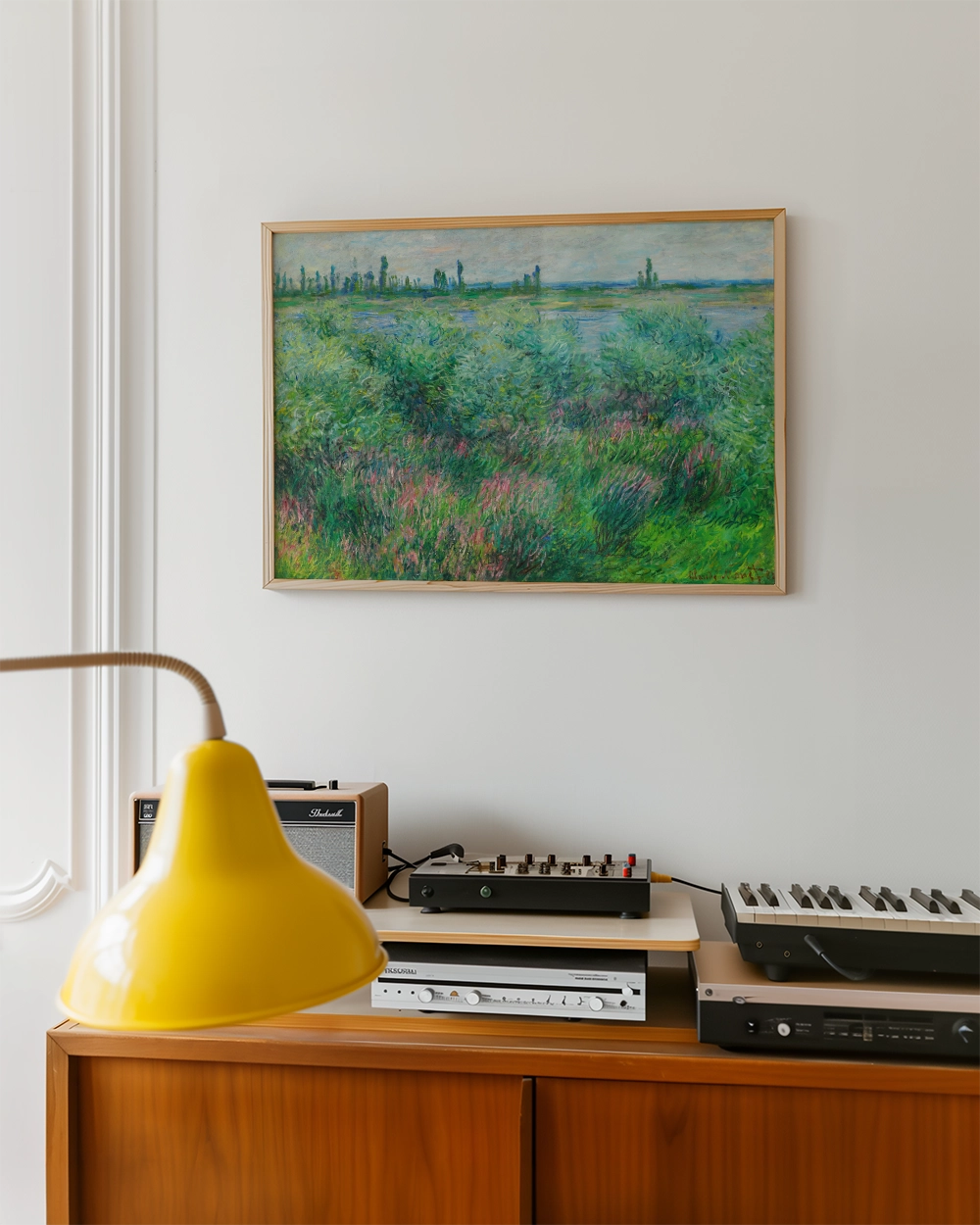 Painting of a lush landscape with greenery and a body of water, framed and displayed on a white wall above a wooden cabinet with a yellow lamp and vintage audio equipment.