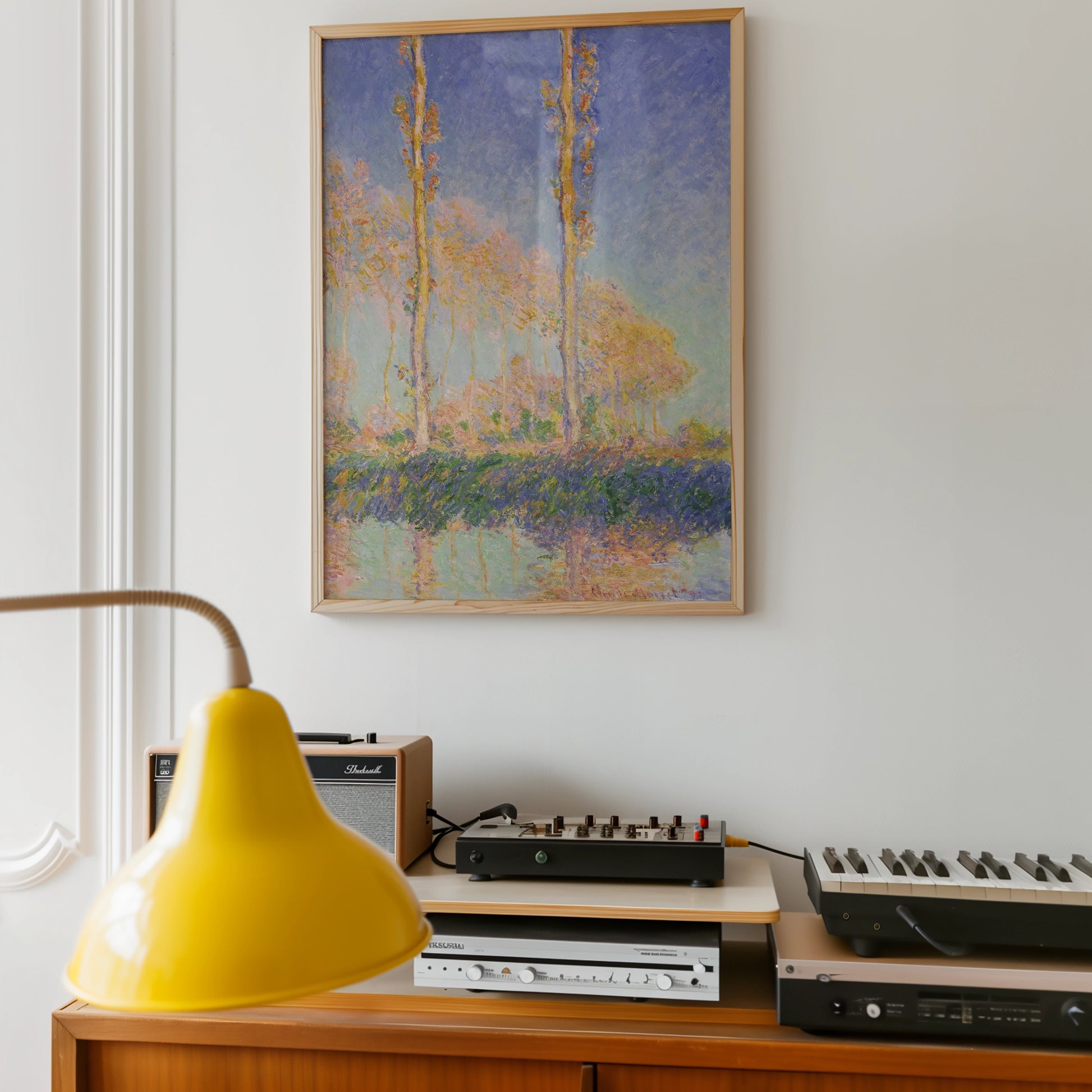 Framed painting of colorful trees reflected in water, displayed on a white wall above retro audio equipment and a yellow lamp.