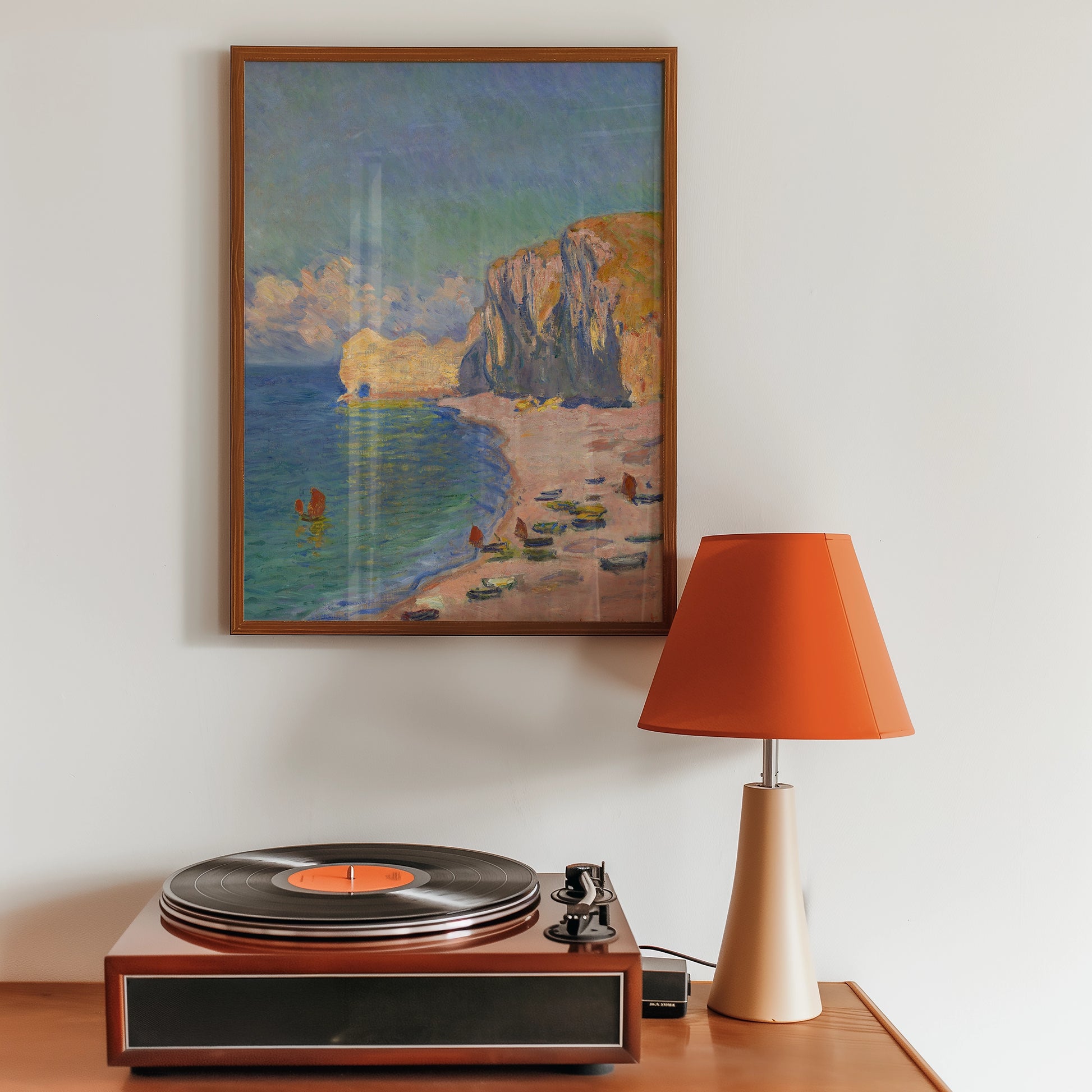 Painting of a coastal scene with cliffs, beach, and boats, framed and displayed above a wooden table featuring a record player and an orange lamp.