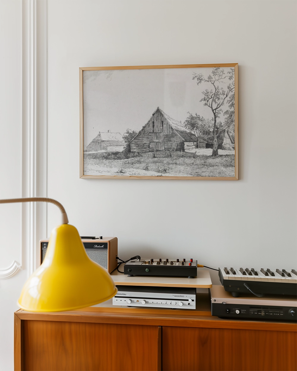 Black and white artwork of rural houses in a wooden frame displayed on a wall above a mid-century modern wooden cabinet with musical equipment and a yellow lamp.