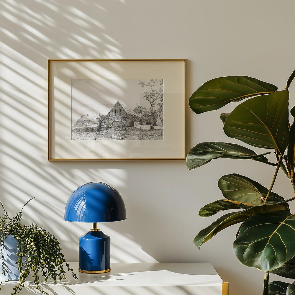 Framed black and white painting of a house surrounded by trees on a wall, with a blue lamp and plant in the foreground.