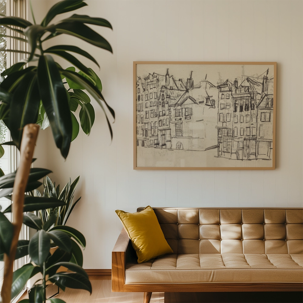 Framed sketch of urban buildings hanging on a wall above a modern tan couch with a yellow pillow, surrounded by greenery.