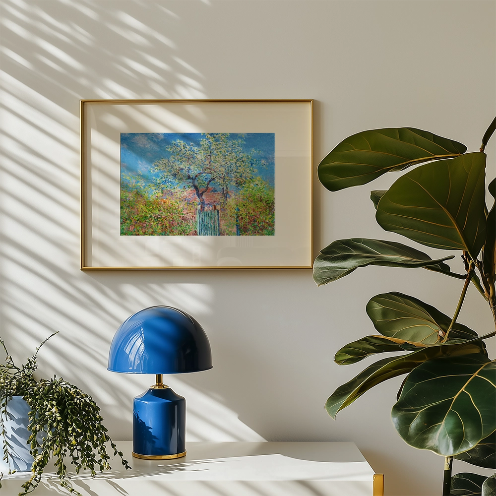 Framed painting of a blooming tree in an impressionistic style, displayed on a light wall with a blue lamp and foliage in the foreground.