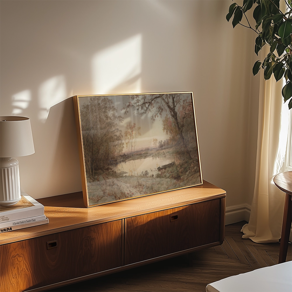 Painting of a serene landscape with trees and water displayed in a stylish frame on a modern wooden table next to a decorative lamp.