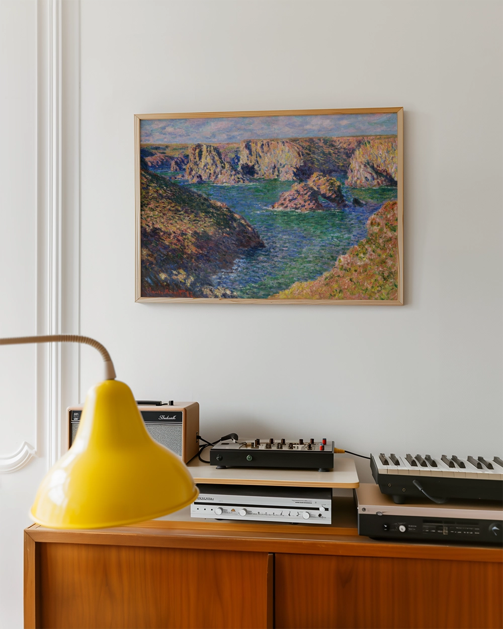 Landscape painting featuring rocky cliffs and a serene body of water, framed and hung on a wall above a wooden console with modern audio equipment and a yellow lamp.