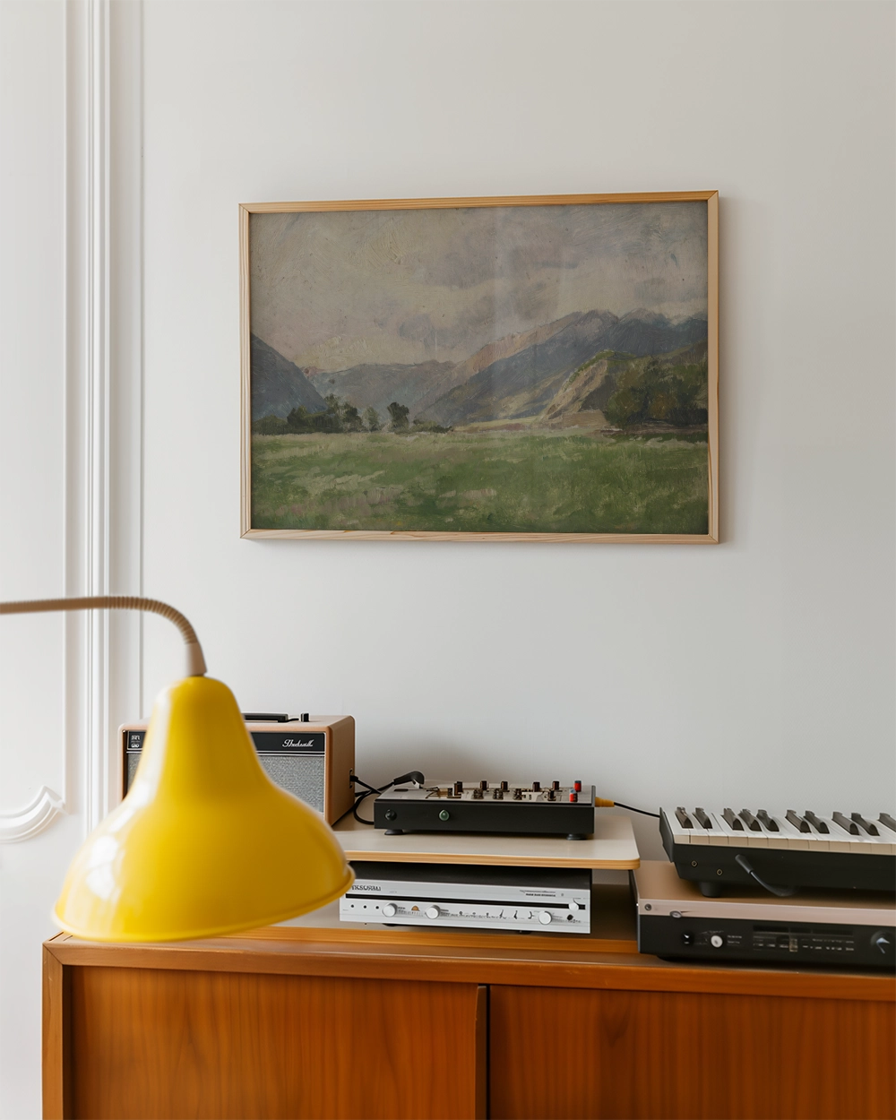 Framed landscape painting of mountains and greenery displayed on a wall above a wooden console with a yellow lamp and audio equipment.