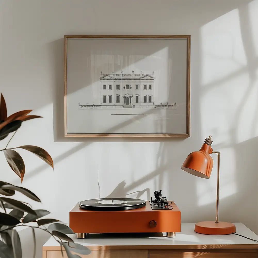 Framed architectural print on a wall above an orange vintage record player and lamp on a wooden surface with green plant leaves in the foreground.