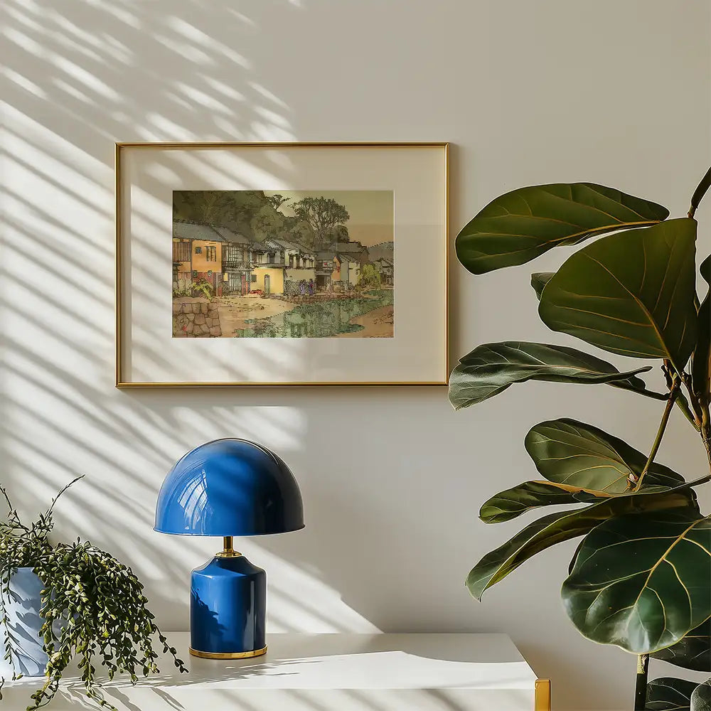Framed painting of a rural scene with houses and a body of water, displayed on a wall beside a blue lamp and a leafy plant.