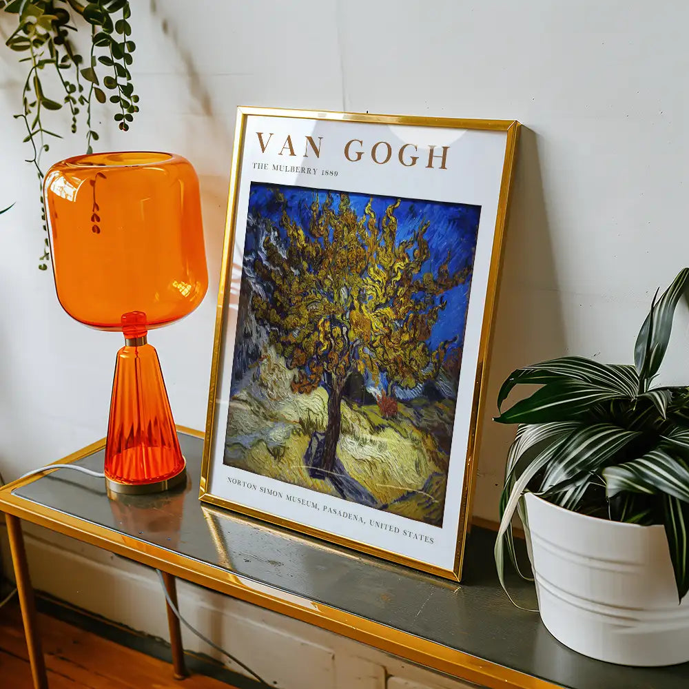 Framed poster of Vincent van Gogh's painting "The Mulberry Tree" (1889) displayed next to an orange table lamp and a potted plant on a stylish table.