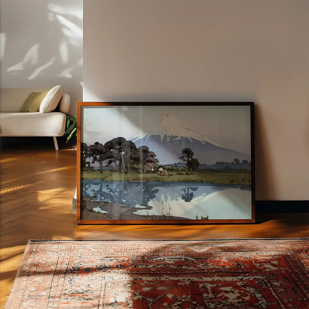 Framed landscape painting featuring a serene mountain scene reflected in a calm body of water, placed on a wooden floor with a decorative rug and living room setting.