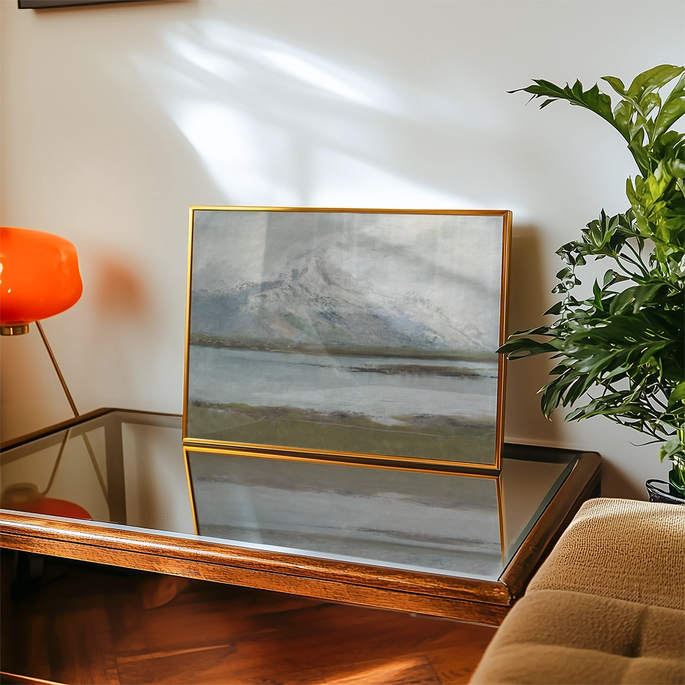 Framed landscape painting with mountains and a lake displayed on a glass table next to a green plant and an orange lamp.
