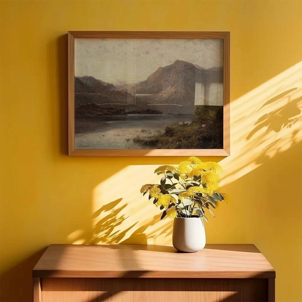 Framed painting of a serene landscape featuring mountains and a lake, displayed on a yellow wall above a wooden table with a vase of yellow flowers.