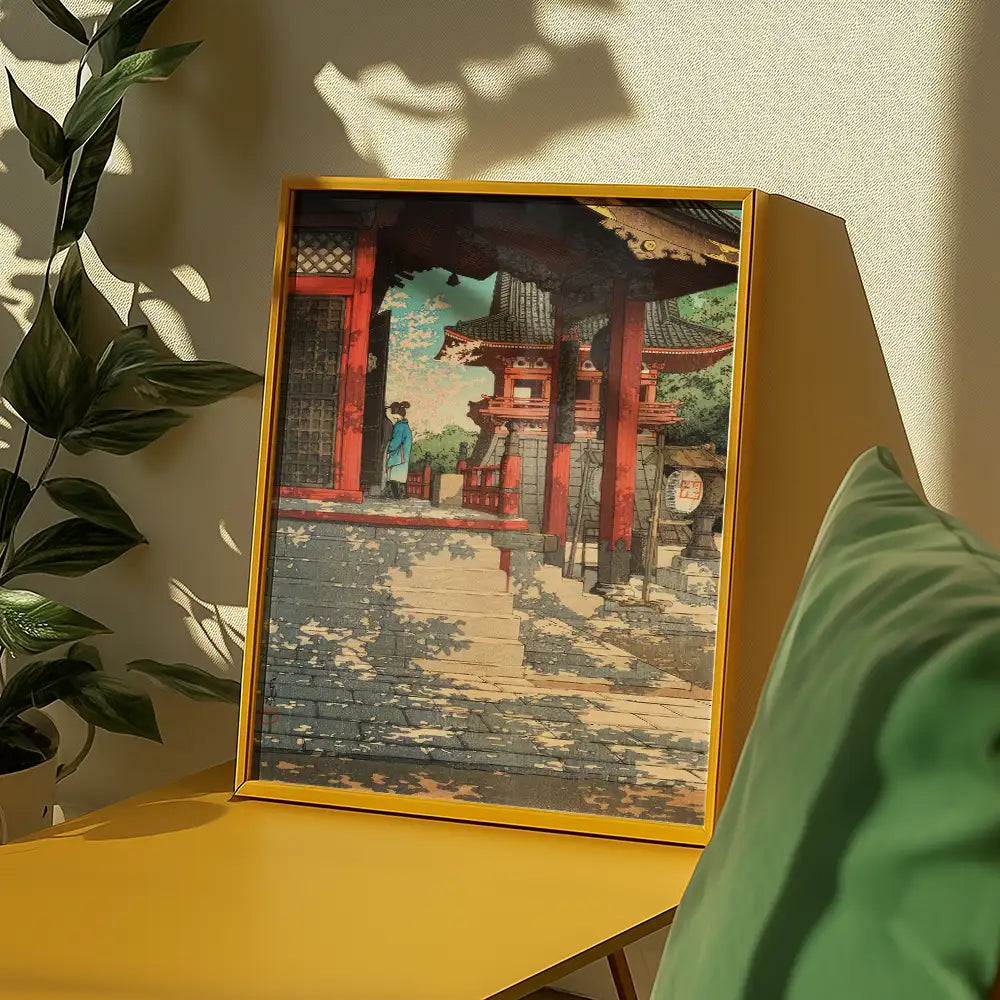Framed painting depicting a traditional Japanese-style building with a figure in a blue garment, set against a backdrop of greenery and shadows, displayed on a yellow table next to a green cushion.
