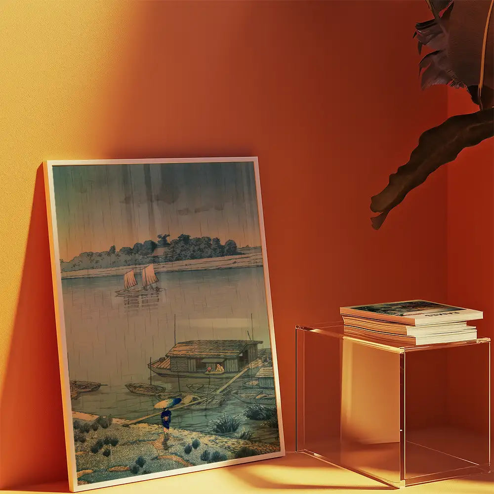 A framed painting of a serene landscape with boats on a river, displayed against an orange wall next to a stack of art books.
