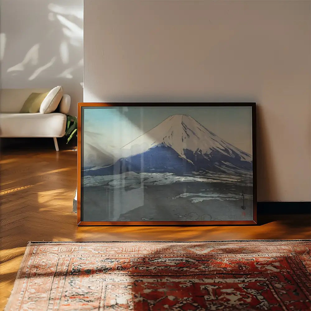A framed painting of a mountain landscape featuring a prominent snow-capped peak, placed on a wooden floor with a cozy interior background.