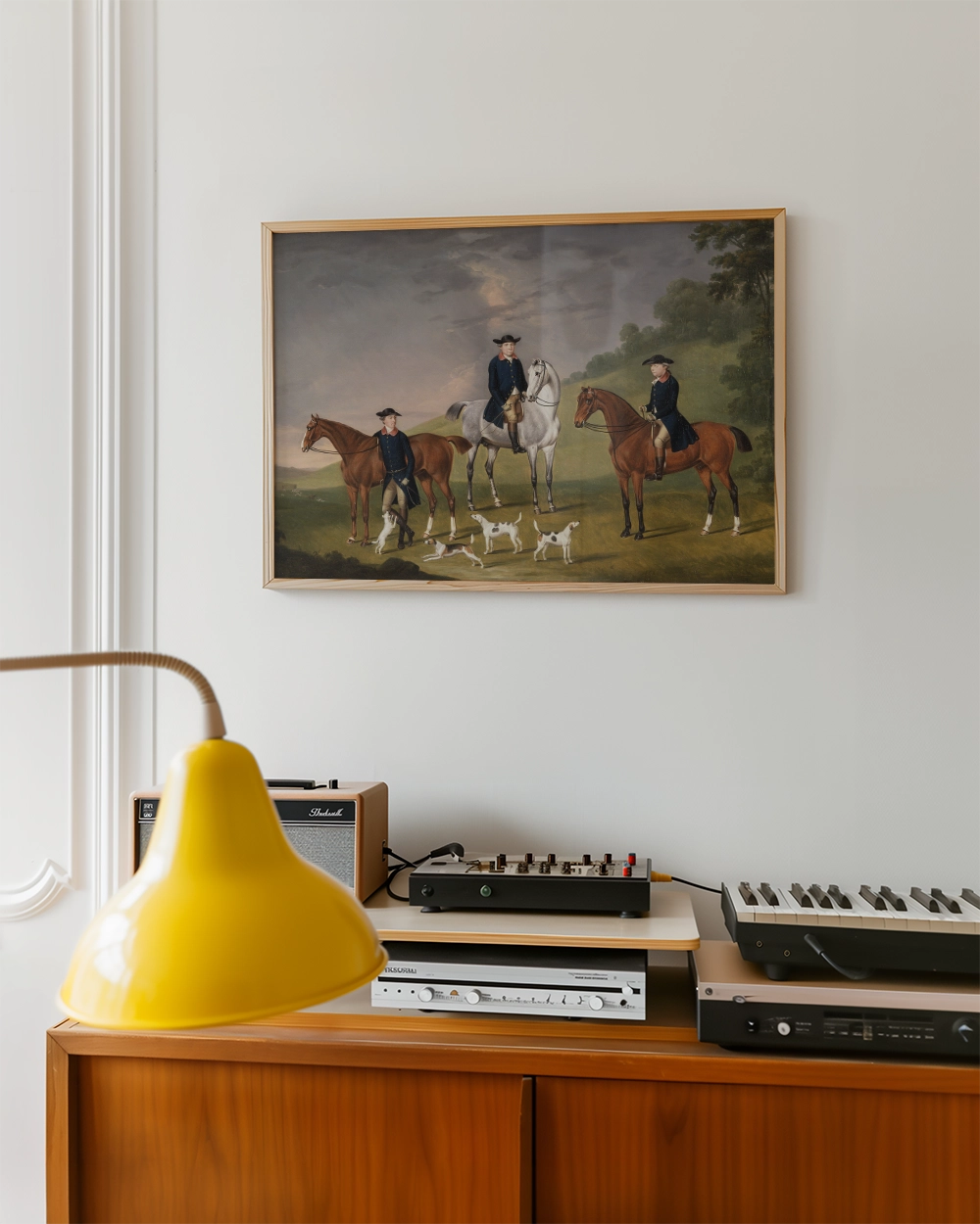 Painting of two horse riders with dogs in a landscape, framed and displayed on a wall above a wooden cabinet with a yellow lamp and audio equipment.