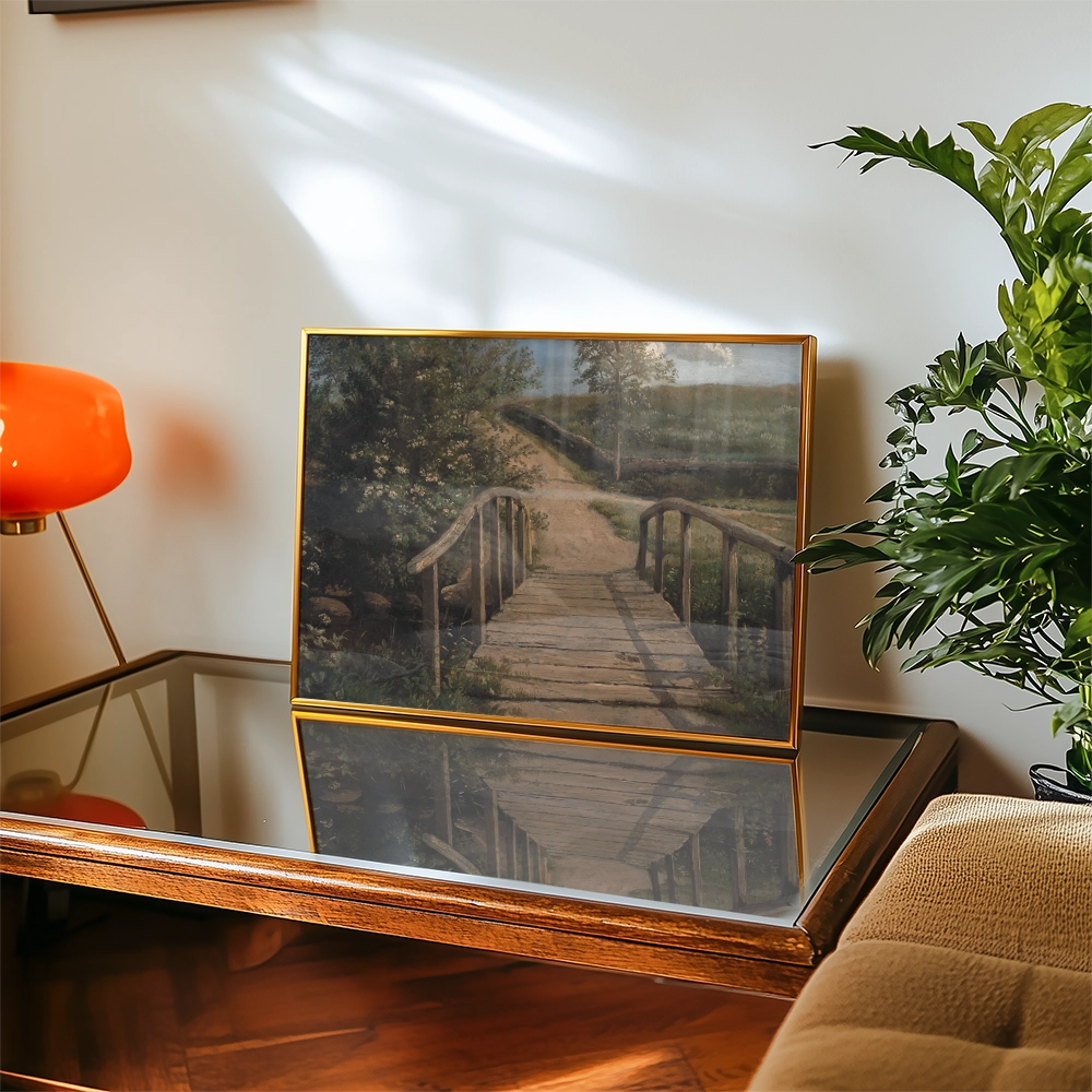 Painting of a serene landscape featuring a wooden bridge over a pathway, framed in gold, displayed on a table with a plant and an orange lamp in the background.
