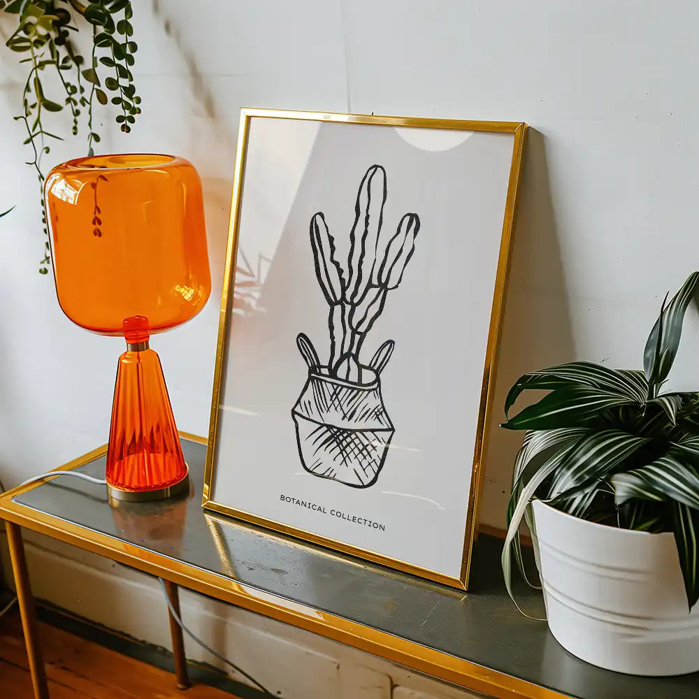 Modern botanical poster in a gold frame featuring a minimalist cactus design, displayed on a stylish table with an orange lamp and green plants.