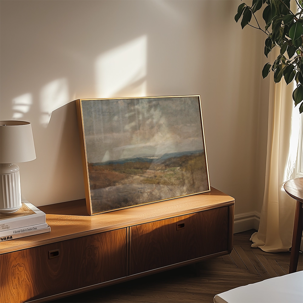 A framed painting of a serene landscape with soft colors displayed on a wooden side table, accompanied by a lamp and a stack of books in a cozy room setting.