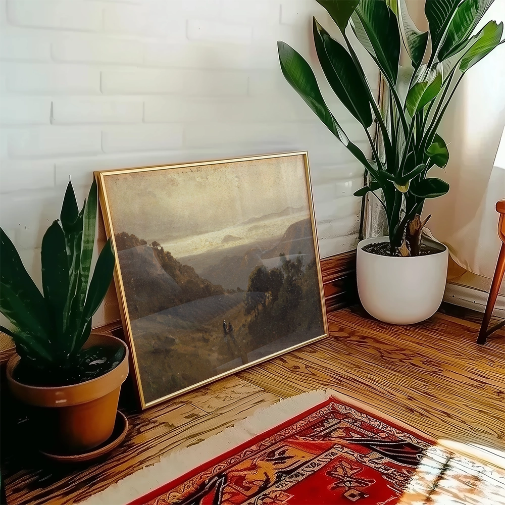Framed landscape painting leaning against a wall next to a decorative plant in a bright interior setting.