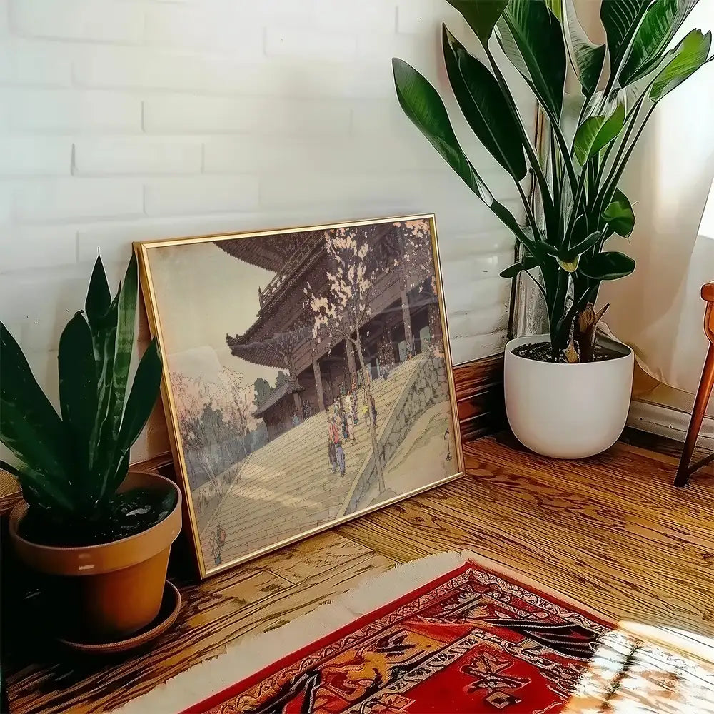 Framed painting of a traditional building with cherry blossom trees, displayed on a wooden floor next to potted plants.