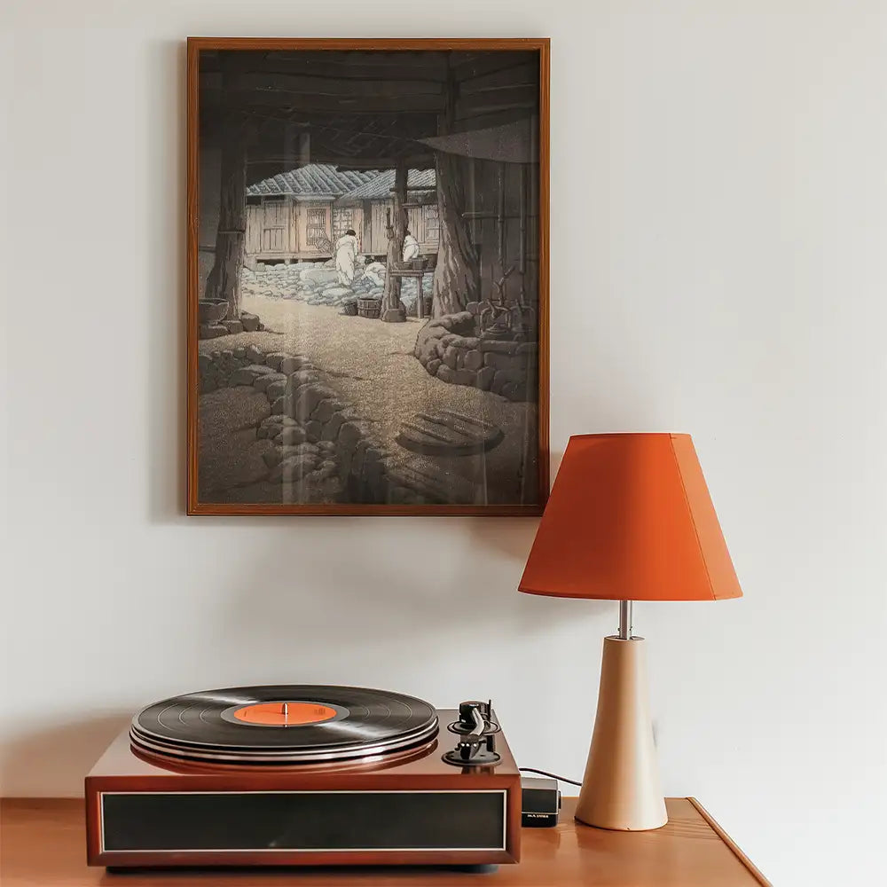 Framed painting depicting a serene traditional scene hung above a vintage record player and a modern lamp with an orange shade.