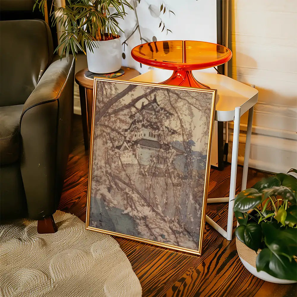 Framed painting featuring a serene landscape with cherry blossom trees and a distant building, placed next to a modern coffee table and greenery in a cozy living space.