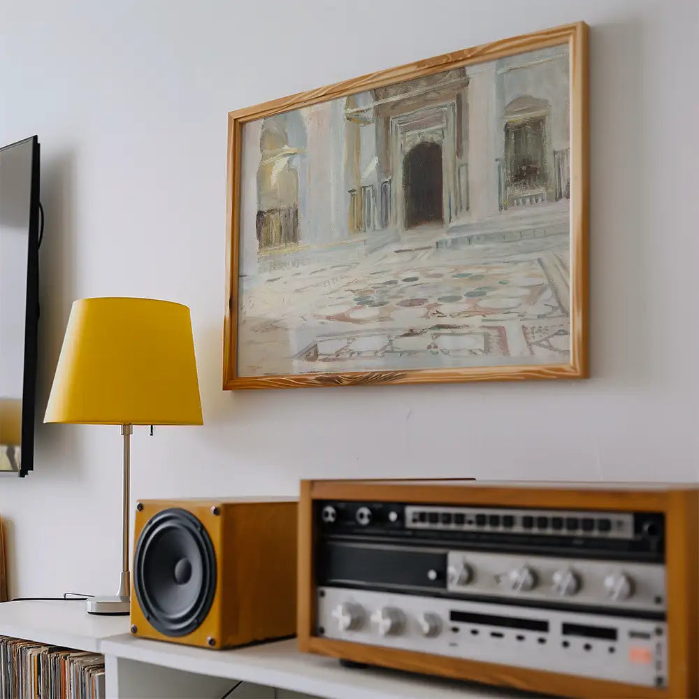 Painting of an architectural scene in a light wooden frame displayed on a wall, with a yellow lamp and vintage audio equipment nearby.
