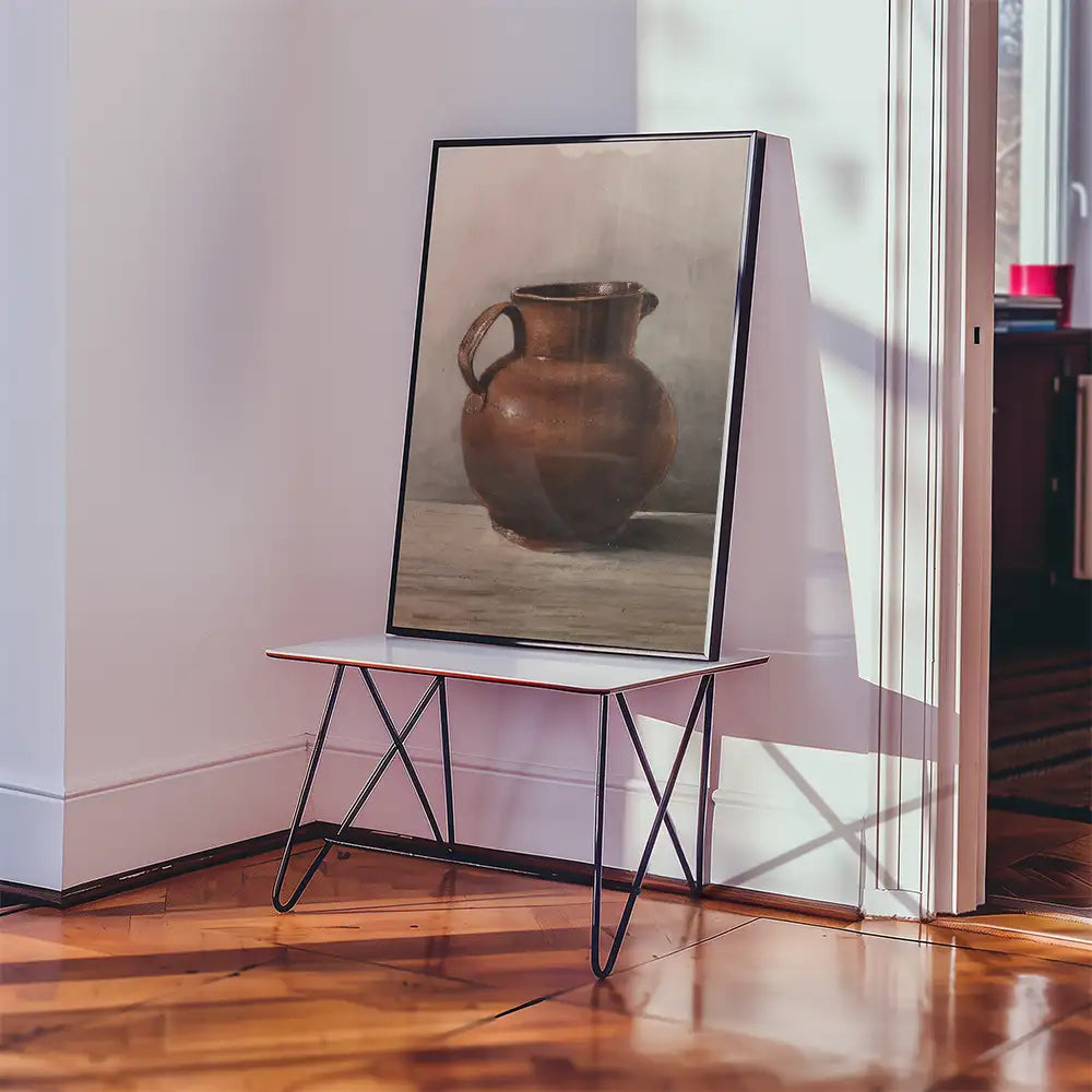 Framed painting of a brown ceramic pitcher displayed on a modern table in a bright room.