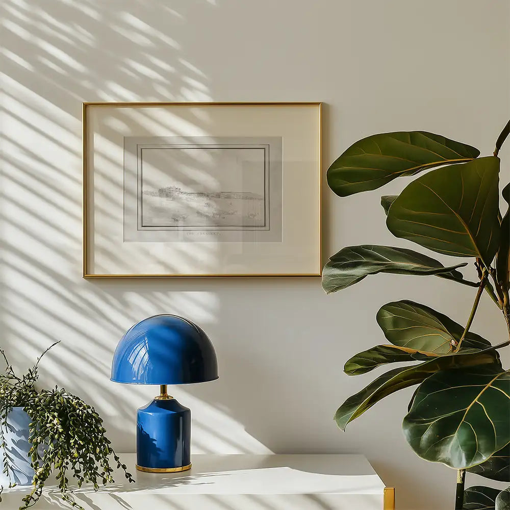 Framed black and white landscape painting hanging on a wall next to a blue lamp and a green plant on a white table