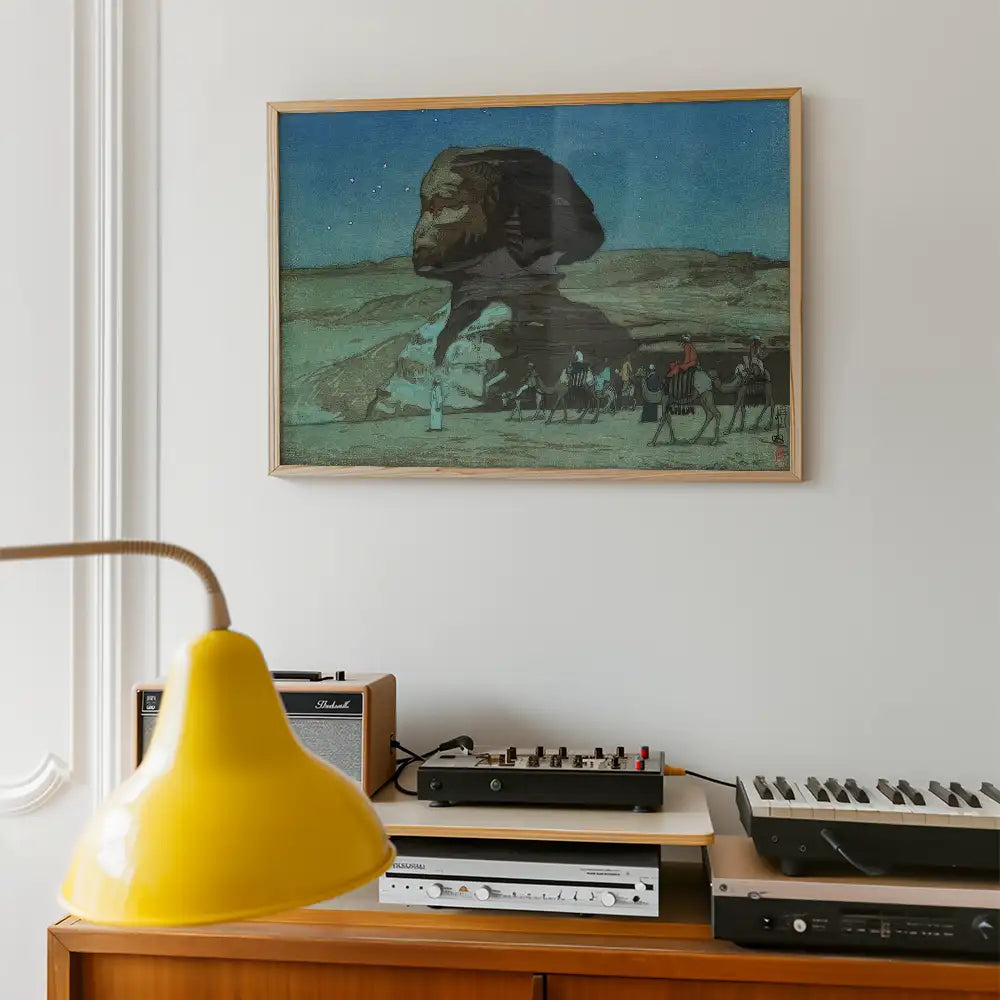 Framed painting of a Sphinx-like figure in a desert landscape, accompanied by a caravan of camels, displayed on a wall above a wooden console with a yellow lamp and audio equipment.