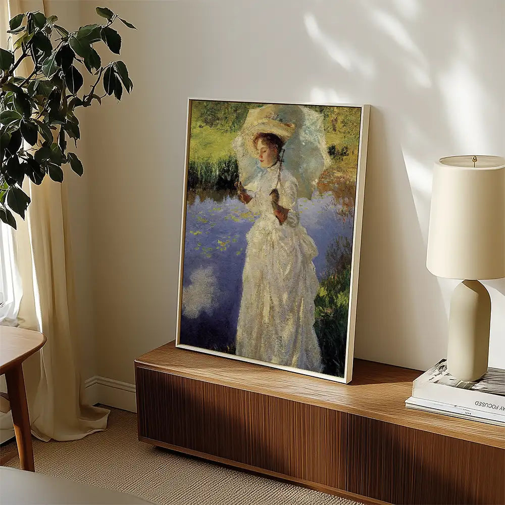 Painting of a woman in a white dress holding an umbrella standing by a pond, framed and displayed on a wooden side table with a lamp and books in a well-lit room.