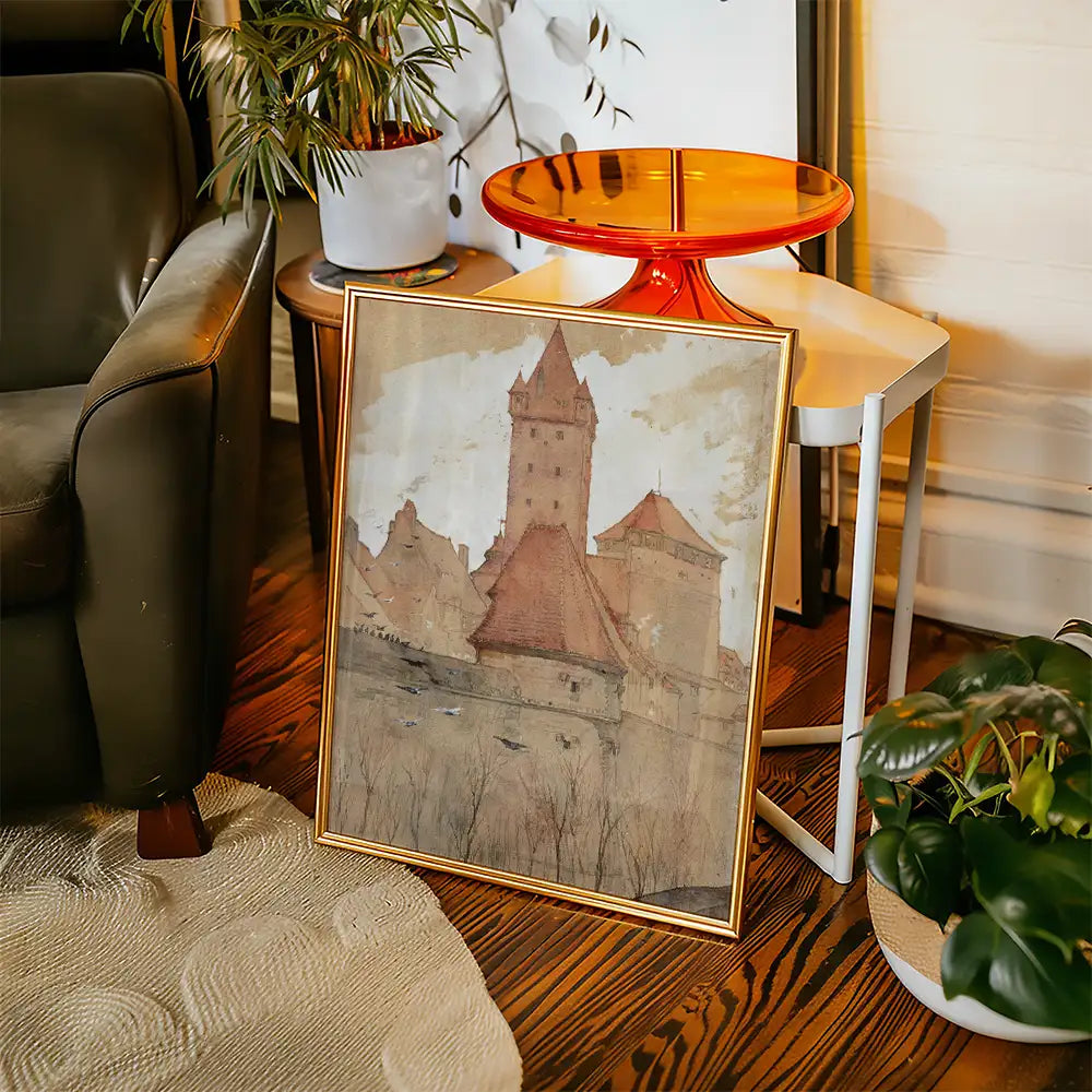 Framed watercolor painting of a castle with red roofs leaning against a modern chair next to a small red side table and a potted plant.