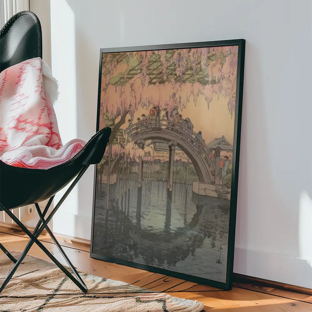 Framed painting of a serene landscape featuring a bridge surrounded by cherry blossom trees, leaning against a wall beside a modern black chair and a patterned rug.