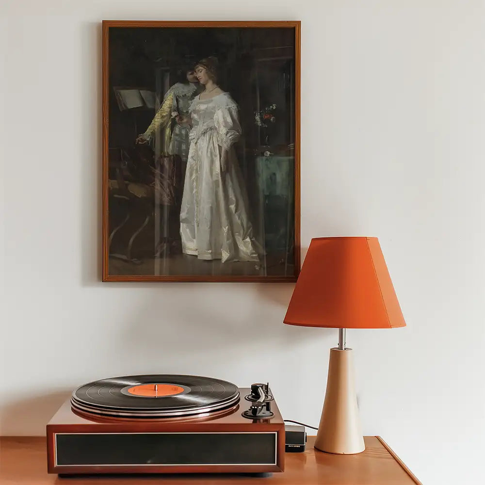 Framed vintage painting of a couple in a romantic setting hanging on a wall above a wooden record player and an orange lamp.