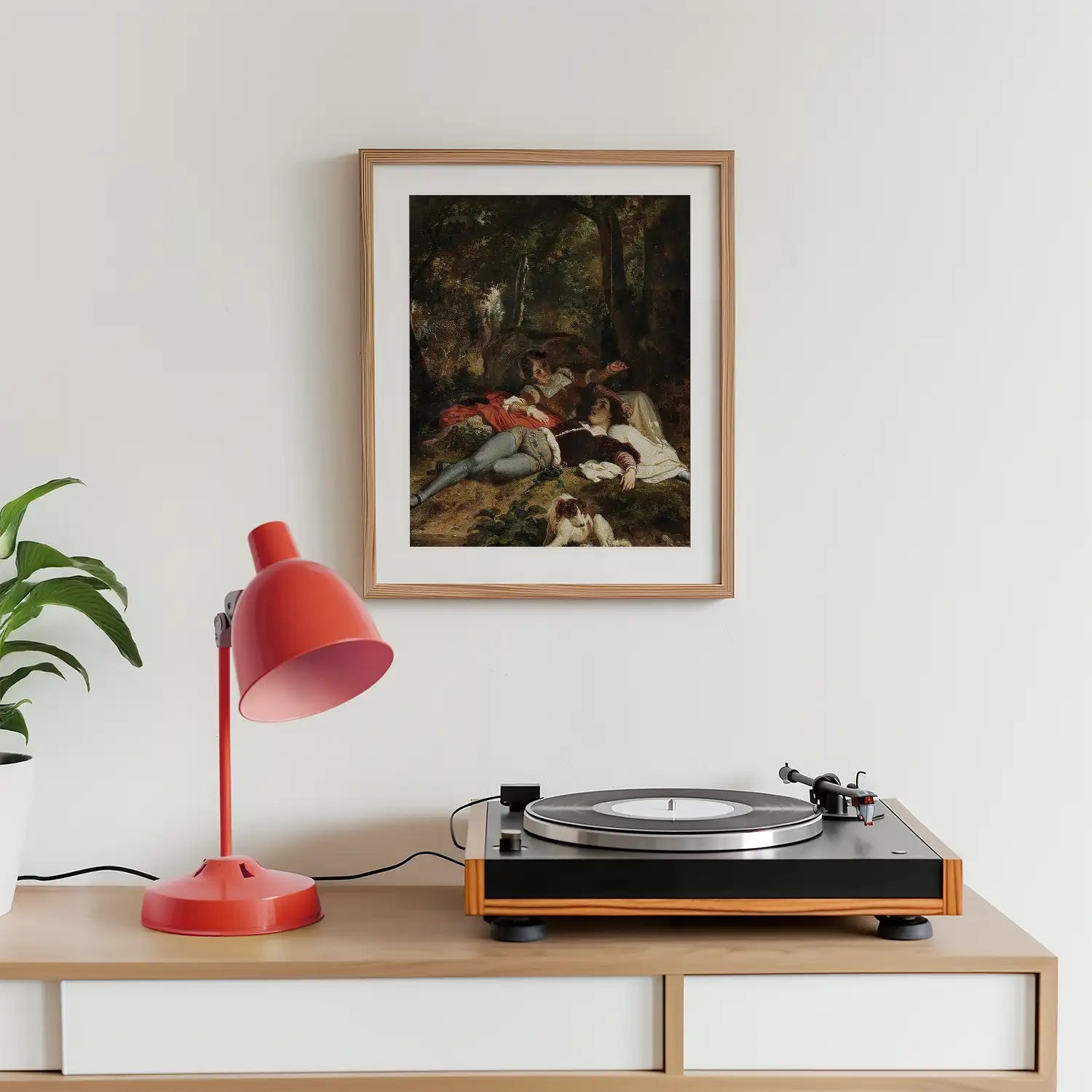 Framed painting of a serene outdoor scene featuring figures resting, displayed above a modern record player and a red desk lamp on a wooden surface.