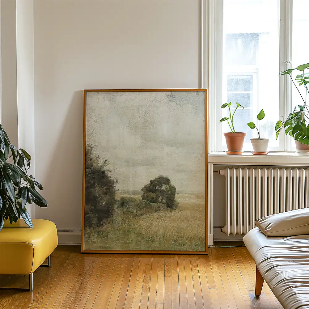 A framed landscape painting depicting a muted rural scene with trees and fields, placed in a modern living room with plants and wooden flooring.