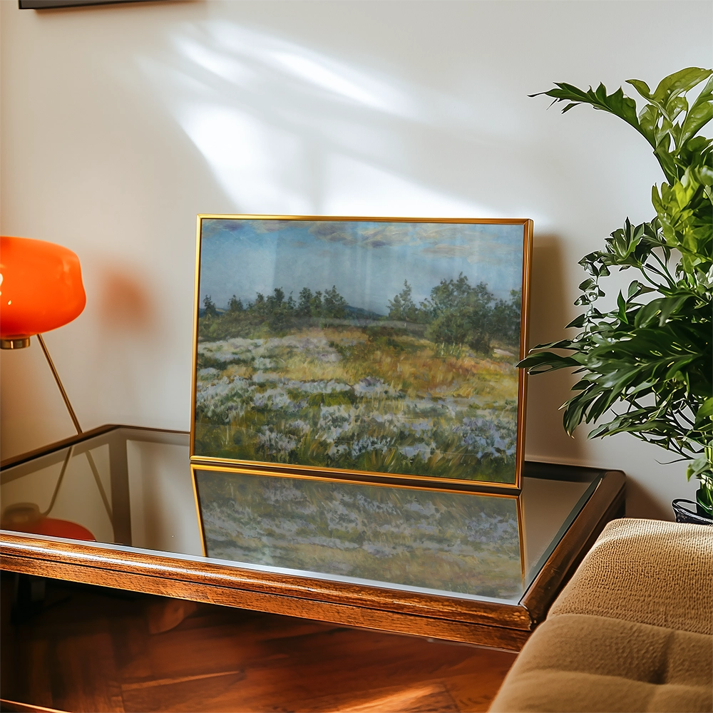 Framed landscape painting featuring a serene field with trees under a blue sky, displayed on a reflective table with a modern lamp and plant in the background.