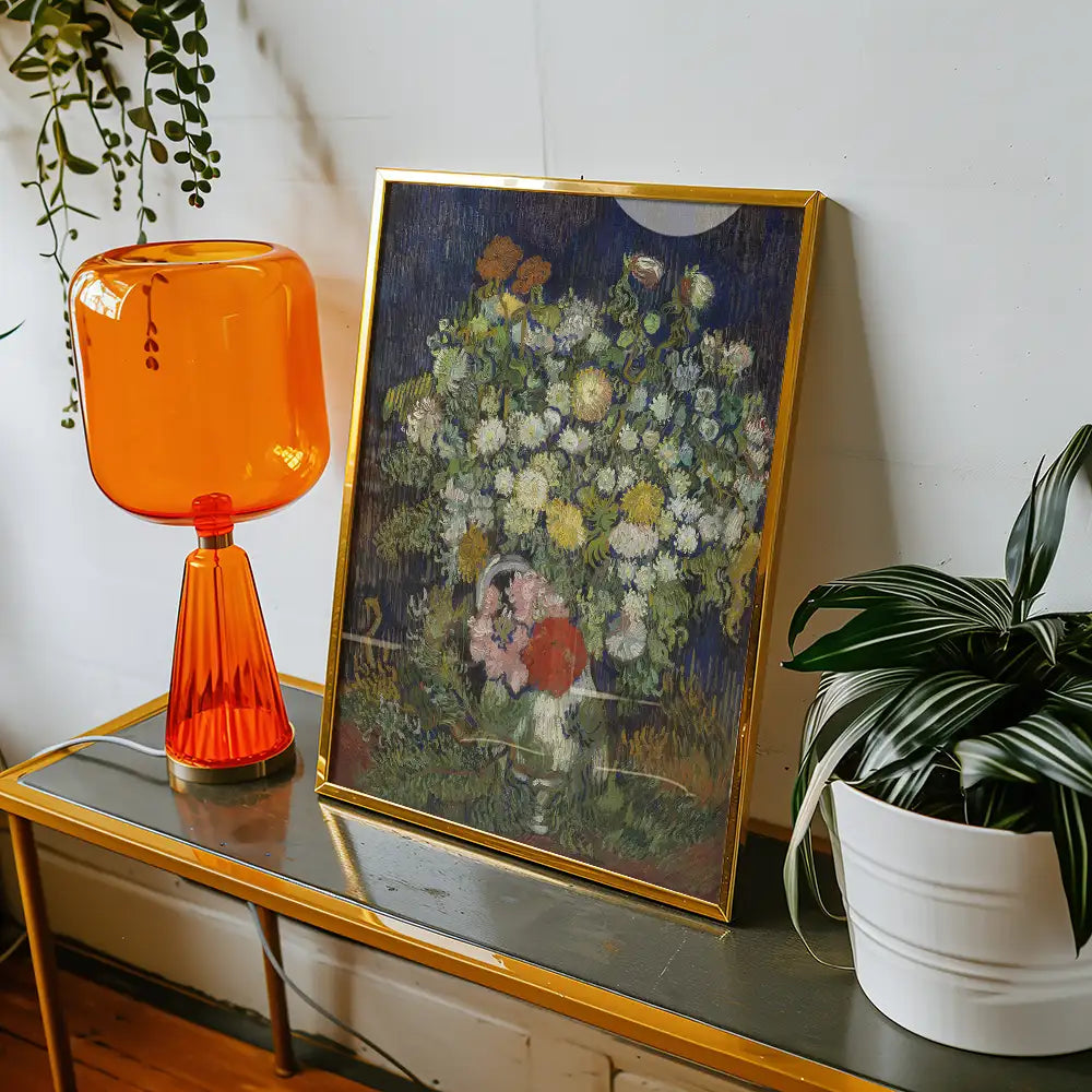 Framed floral painting on a table beside an orange lamp and a potted plant.