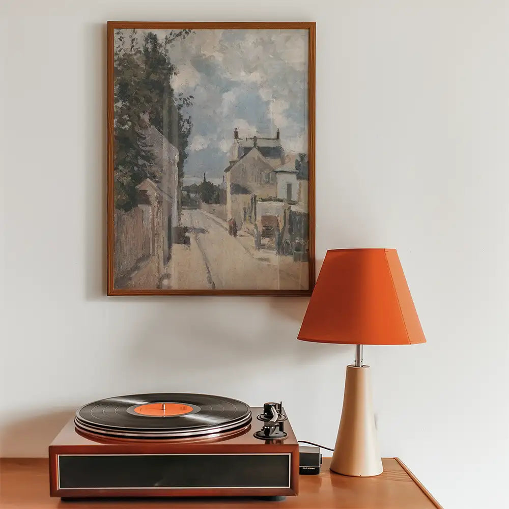 Painting of a street scene in a wooden frame displayed on a wall above a retro record player and an orange lampshade on a wooden table.