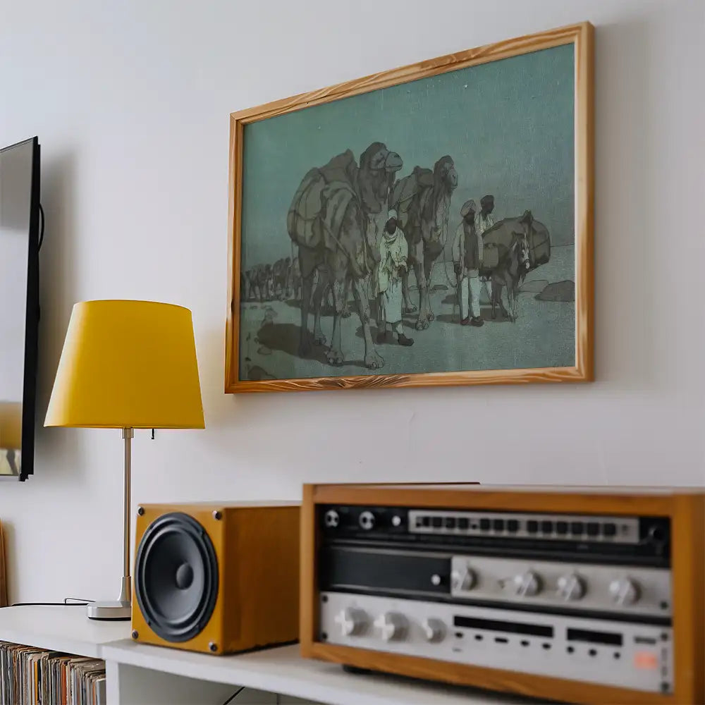 Framed painting of camels and travelers in a muted color palette, displayed on a wall with a yellow lamp and vintage audio equipment nearby.