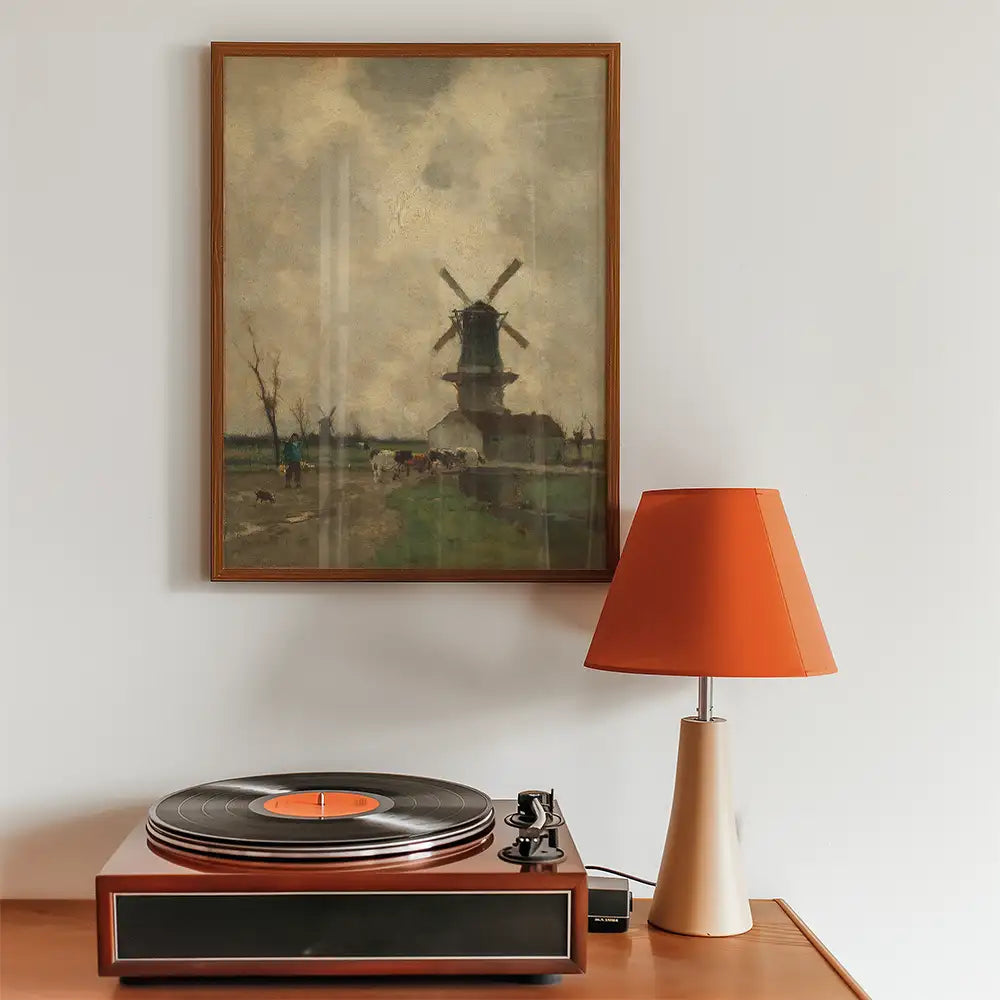 Vintage painting of a windmill hanging above a record player and a modern orange lamp on a wooden surface.